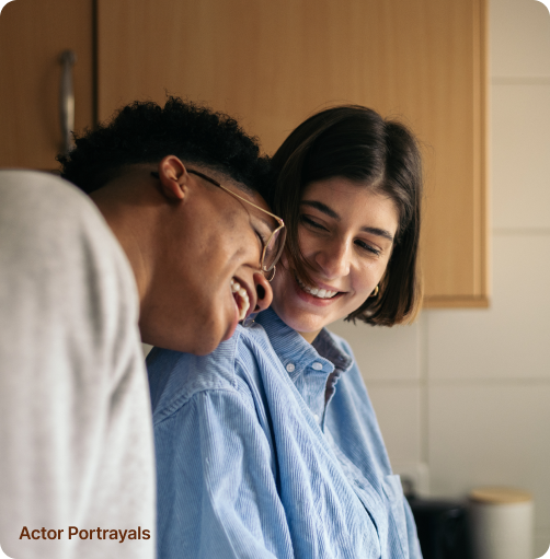A couple standing in the kitchen, with the man leaning over towards the woman, both smiling happily, suggesting a moment of affection and warmth shared between them.