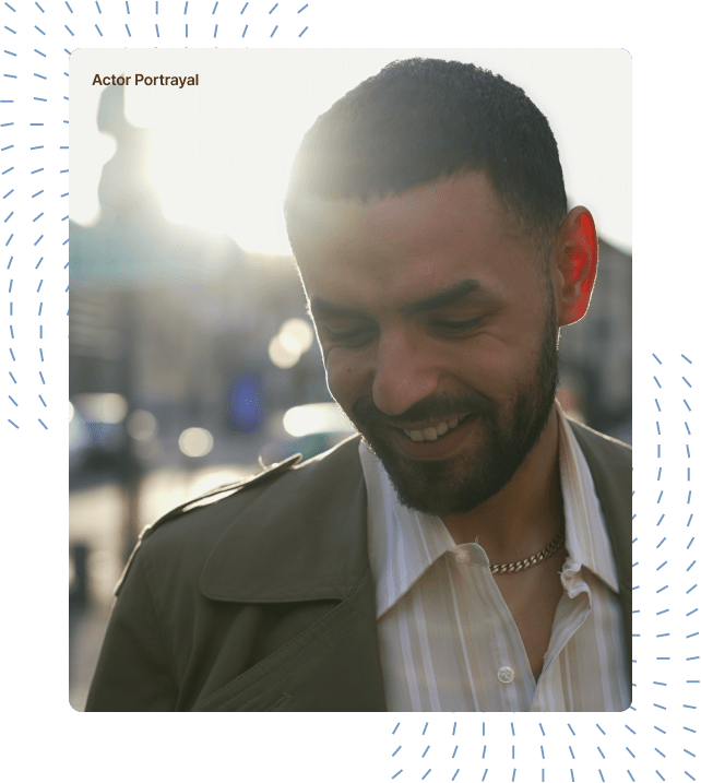 A man wearing an olive jacket standing outdoors, smiling with his gaze directed downward, while sunlight filters through the background, casting a warm glow around him.