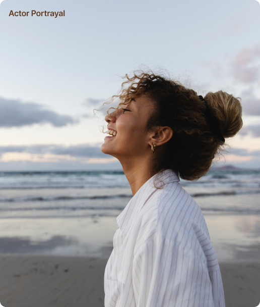 Joyful woman enjoying a peaceful moment at the beach with a soft smile, white shirt billowing in the breeze.