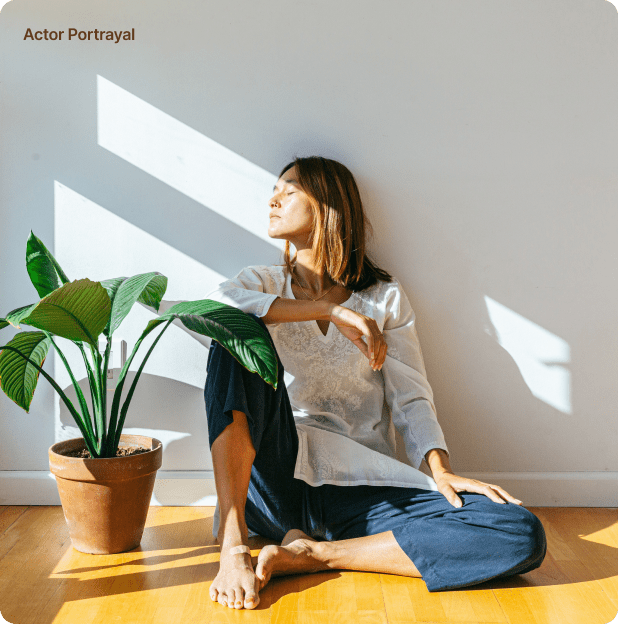 Woman in contemplative pose by a window with sunlight casting shadows, near a lush potted plant.